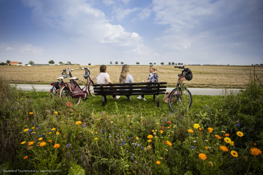 Drei Frauen sitzen auf einer Bank umgeben von Feldern. Sie unterhalten sich. Neben der Bank stehen Fahrr&auml;der. Foto: Sauerland Tourismus e.V./ Foto: Sabrina Voss (sabrinity)